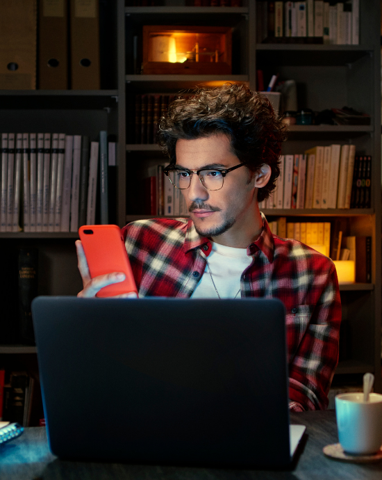 man met bril achter laptop met mobiel in zijn hand